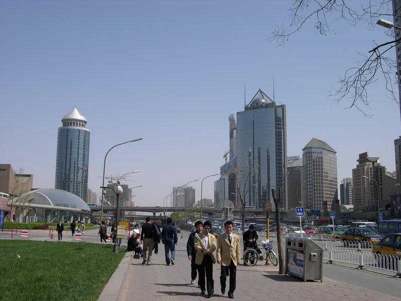 Beijing street scene in bright sunshine with glass towers, an elevated road and people walking along a wide pavement