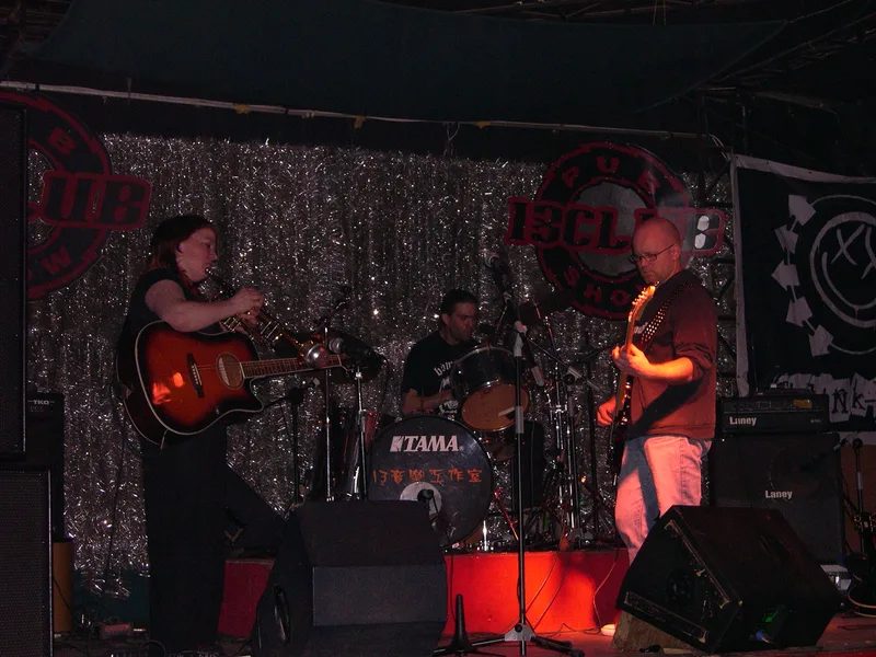 JEF on the 13 Club stage in front of a silver tinsel backdrop: Katy with acoustic guitar, Pez on drums and Kjell on bass