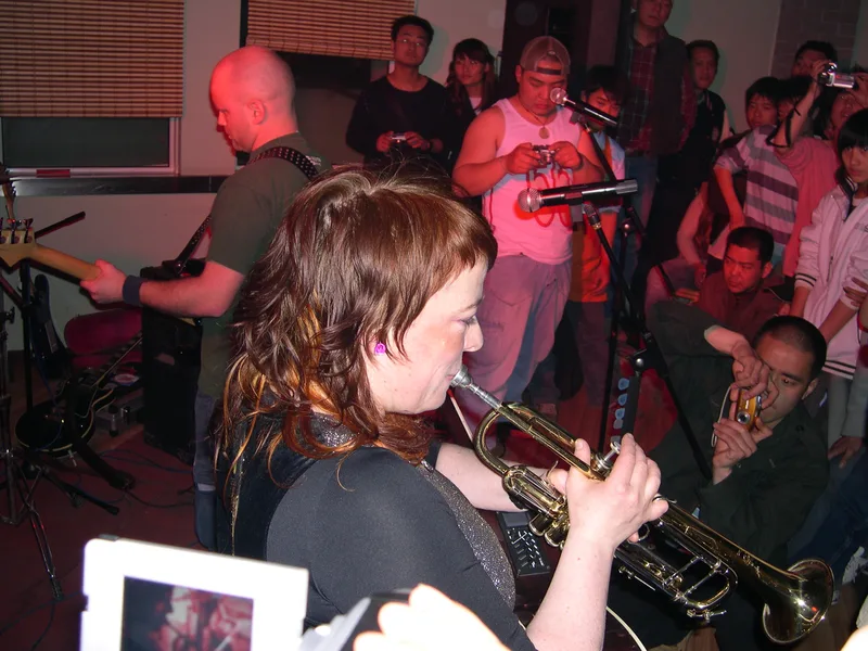 Katy playing trumpet with an acoustic guitar slung on her back, the Xinxiang crowd a metre behind her