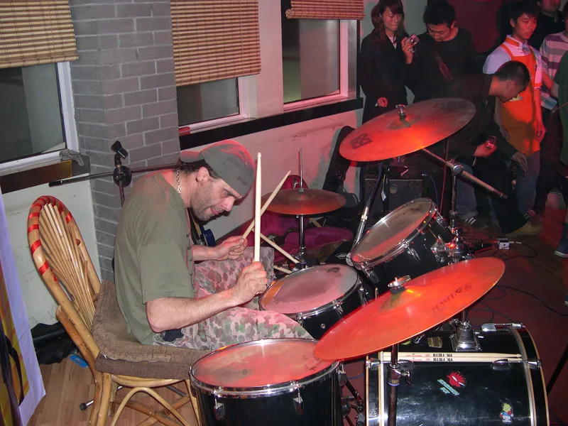 Pez behind a Yamaha drum kit in front of an orange banner, sitting on an ordinary wooden restaurant chair