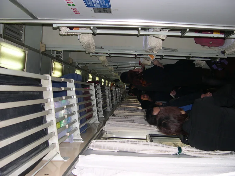 A narrow corridor of a Chinese sleeper train with stacked bunks and passengers resting