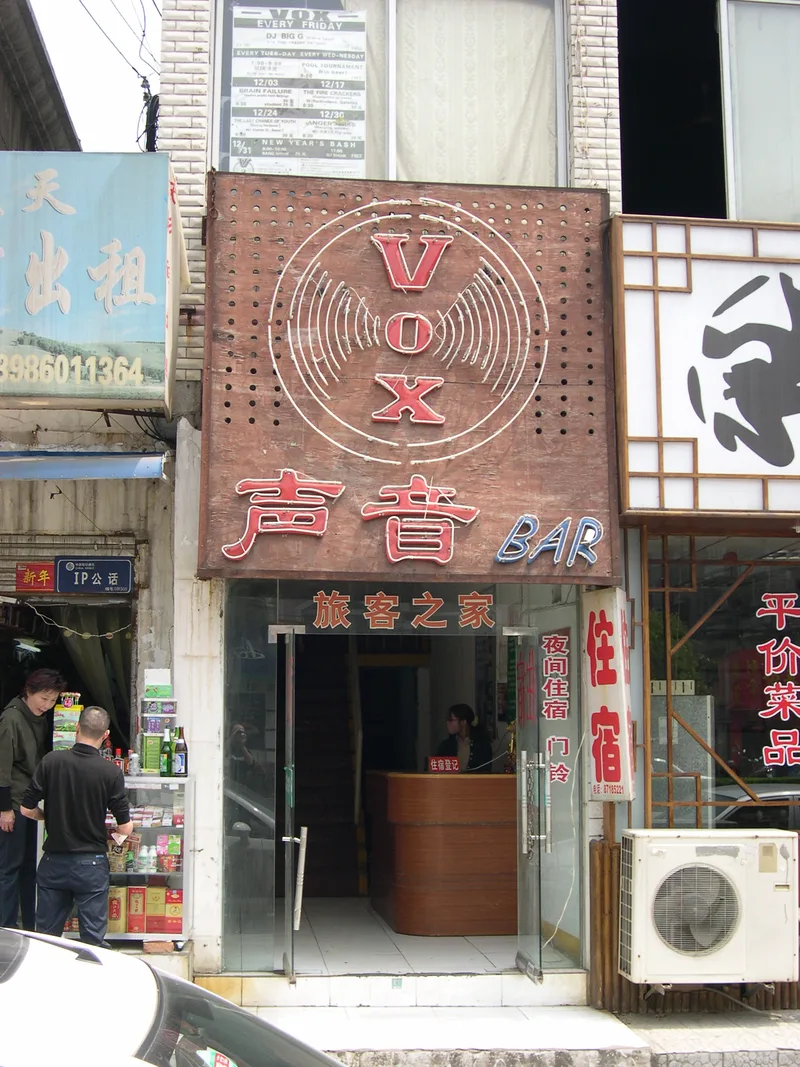 The rusted metal VOX BAR sign with neon tubes, mounted on the street outside the club in Wuhan