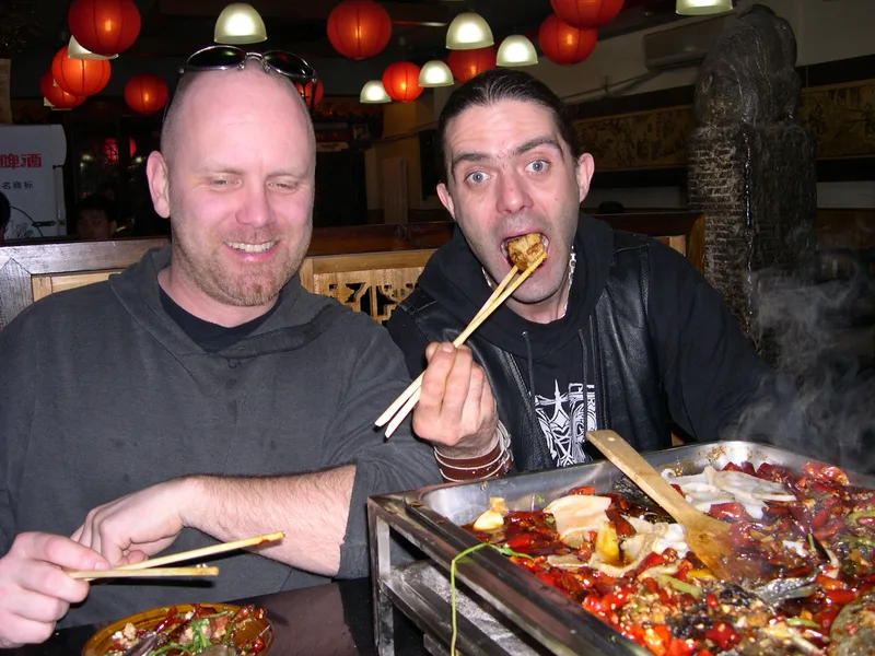 Kjell and Pez at a restaurant table in Beijing eating from a steaming tray of chilli hotpot with chopsticks, red lanterns above
